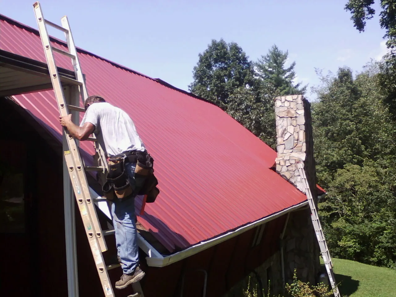 Roofer on a ladder beside a red metal roof and stone chimney — Soffit & Fascia Repair by Roofing Brothers in Farmingdale